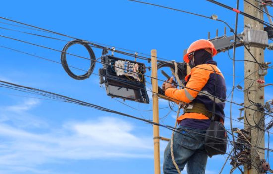 Technician on wooden ladder using smartphone to check data numbers of cable lines while installing fiber optic system in internet splitter box on electric pole against blue sky